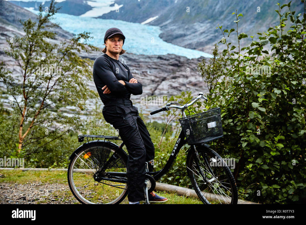 Young man in black dresses on bicycle, with black cap, Svartisen glacier on background Stock Photo