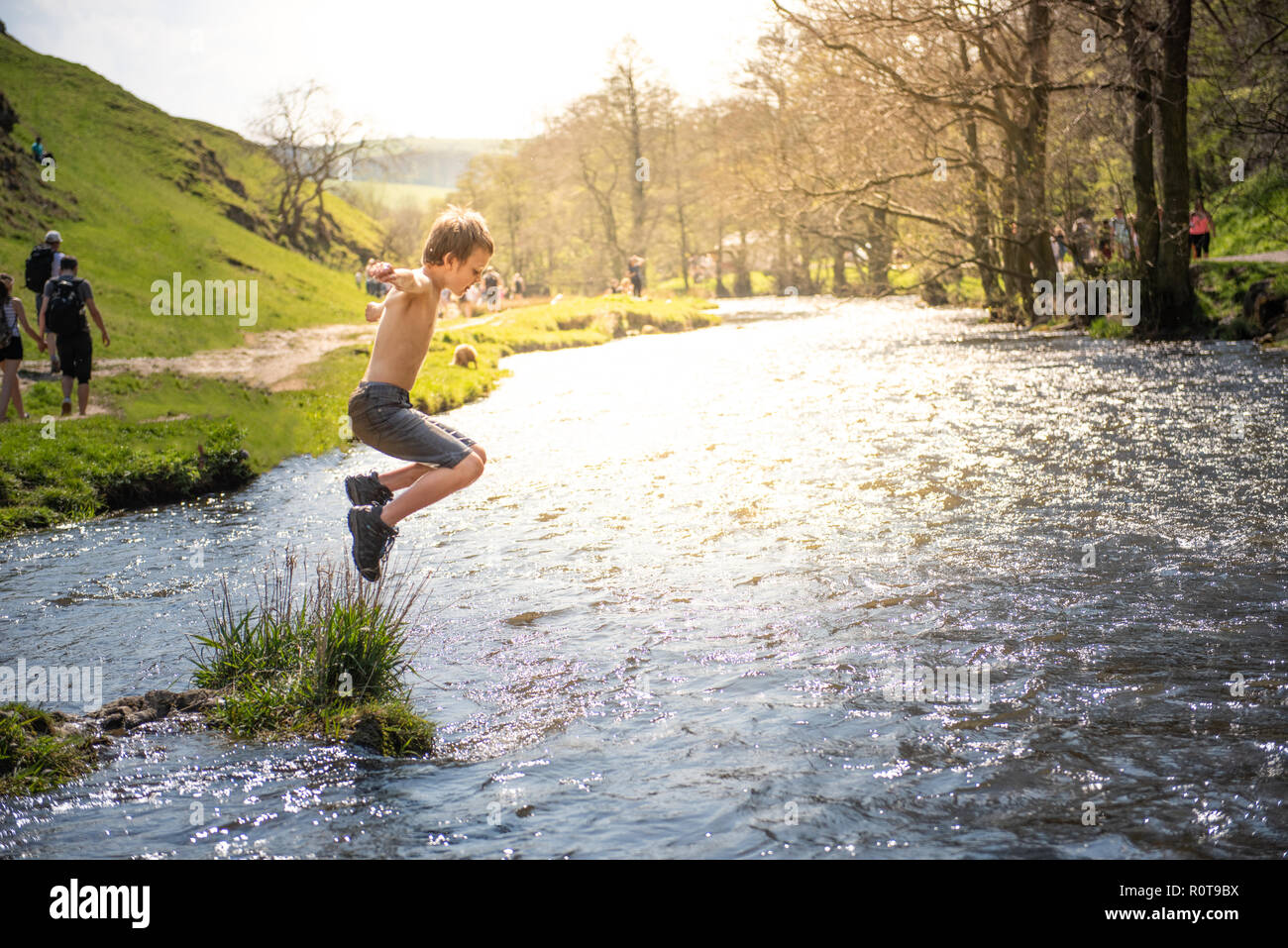 A little boy with ADHD, Autism, Aspergers Syndrome jumps into the river Dove at Dovedale on a hot summers day in denim shorts and hiking boots Stock Photo