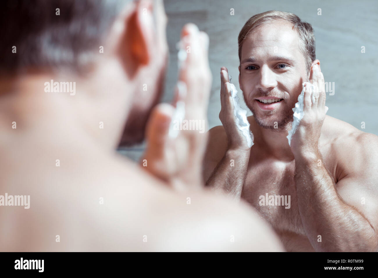 Cheerful blue-eyed man smiling while looking into mirror and shaving face Stock Photo
