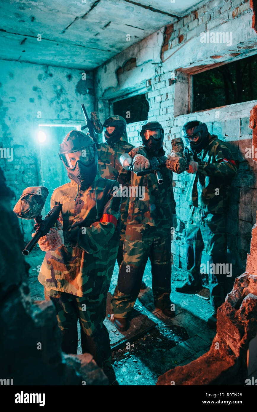 paintball team in uniform and protective masks aiming by paintball guns at camera in abandoned