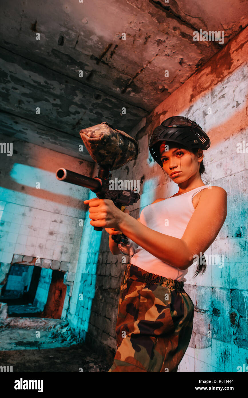 attractive young female paintballer in goggle mask above head aiming by paintball marker in abandoned building Stock Photo