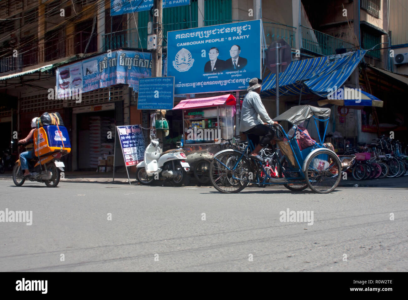 A cyclo & a motorcycle delivery man are riding on a street dominated by a Cambodian People's party campaign sign in Phnom Penh, Cambodia. Stock Photo