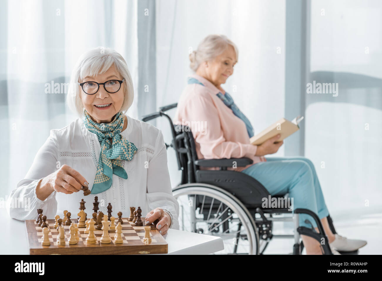 senior woman playing chess at table while another sitting in wheelchair and reading book Stock Photo