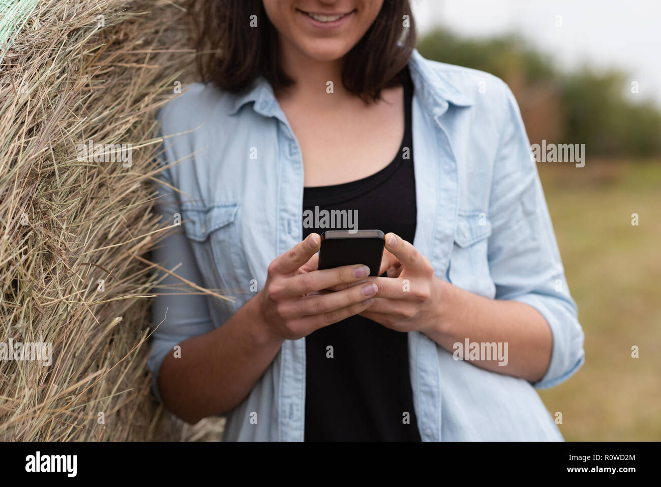 Woman using mobile phone while leaning on hay bale Stock Photo