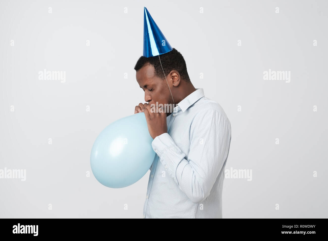 Funny young african guy inflating the balloon wearing blue party hat. Stock Photo