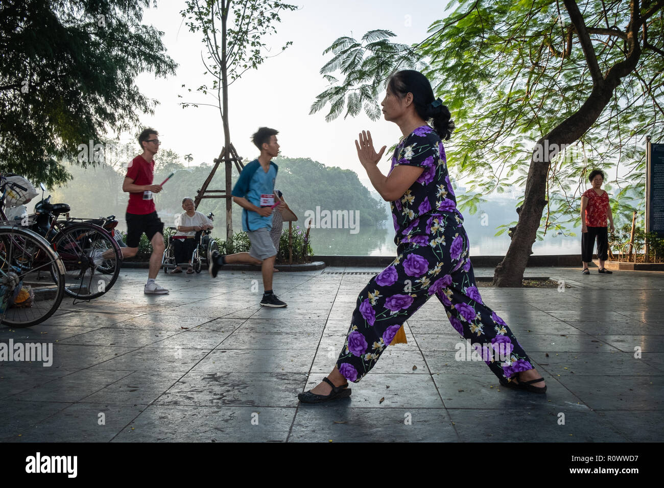 A woman does morning exercise as two men jog past next to Hoan Kim Lake, Hanoi, Vietnam. Stock Photo