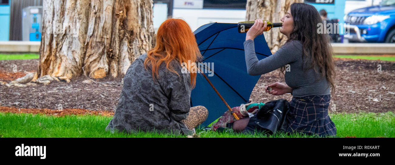 Two young female adults sitting and drinking from a beer bottle in Hyde Park Sydney NSW Australia. Stock Photo