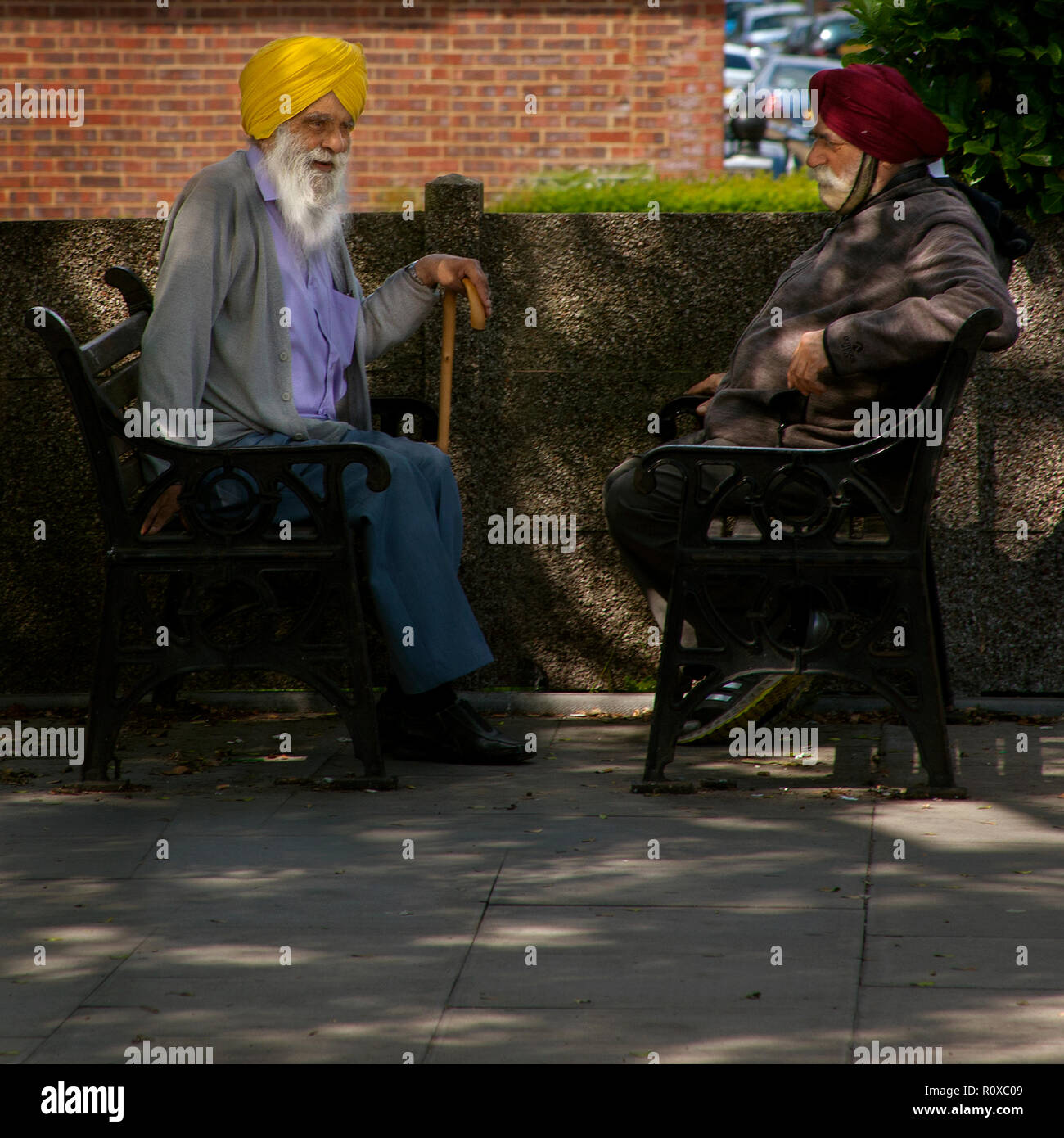 two elderly sikh man wearing yellow and red turbans sitting opposite each other on public benches Southall london Stock Photo