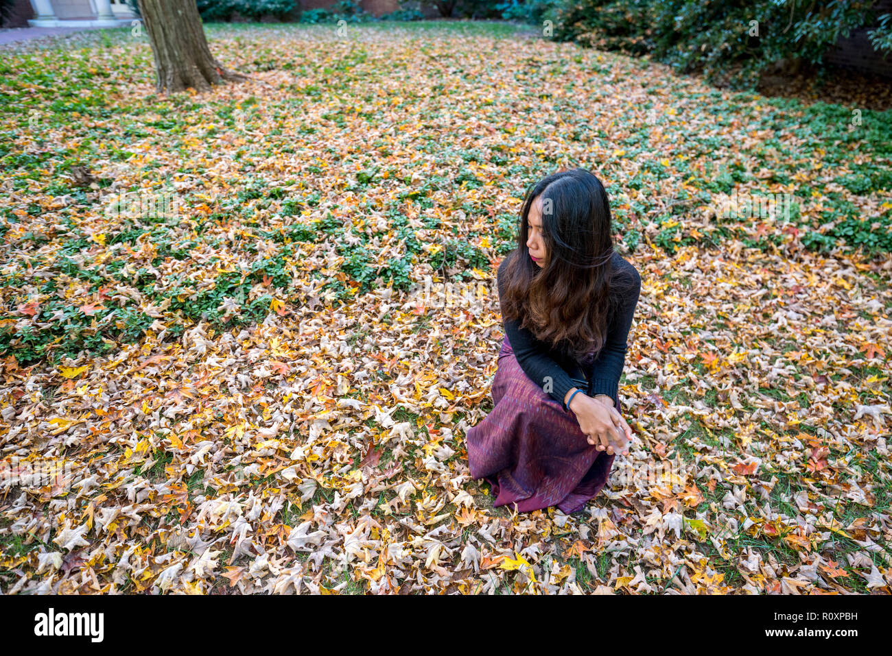 Thai woman kneeling on ground surrounded by leaves in the fall looking to the side Stock Photo