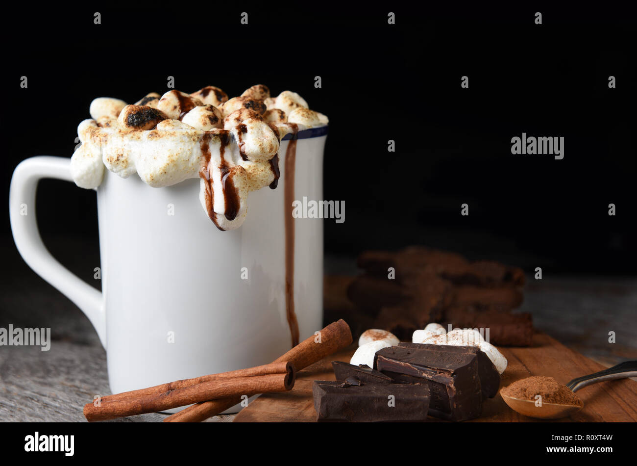 Closeup of a mug of hot cocoa with toasted marshmallows with cookies, chocolate chunks, cinnamon sticks, against a dark background. Stock Photo
