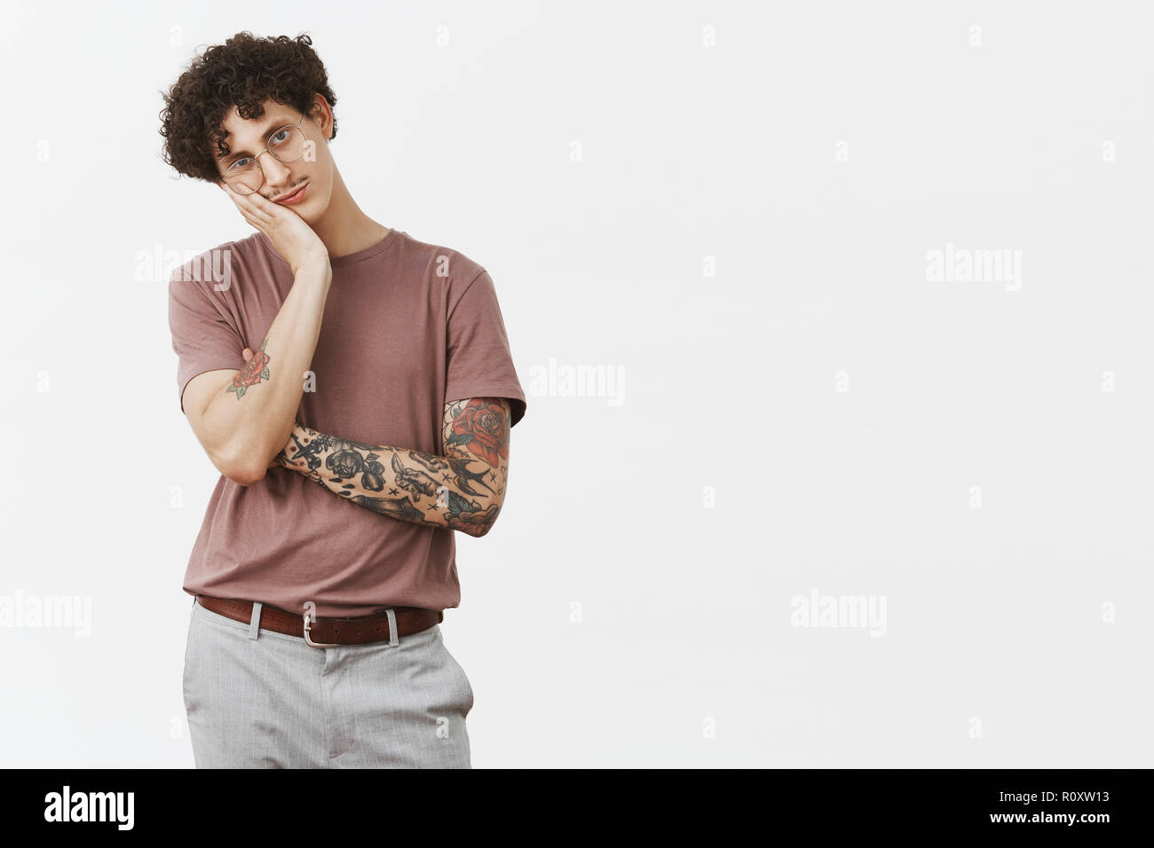 Guy hates sitting at home without action. Portrait of bored handsome and stylish urban young male with moustache, tattooed arm and curly hair leaning on palm indifferent and gloomy Stock Photo
