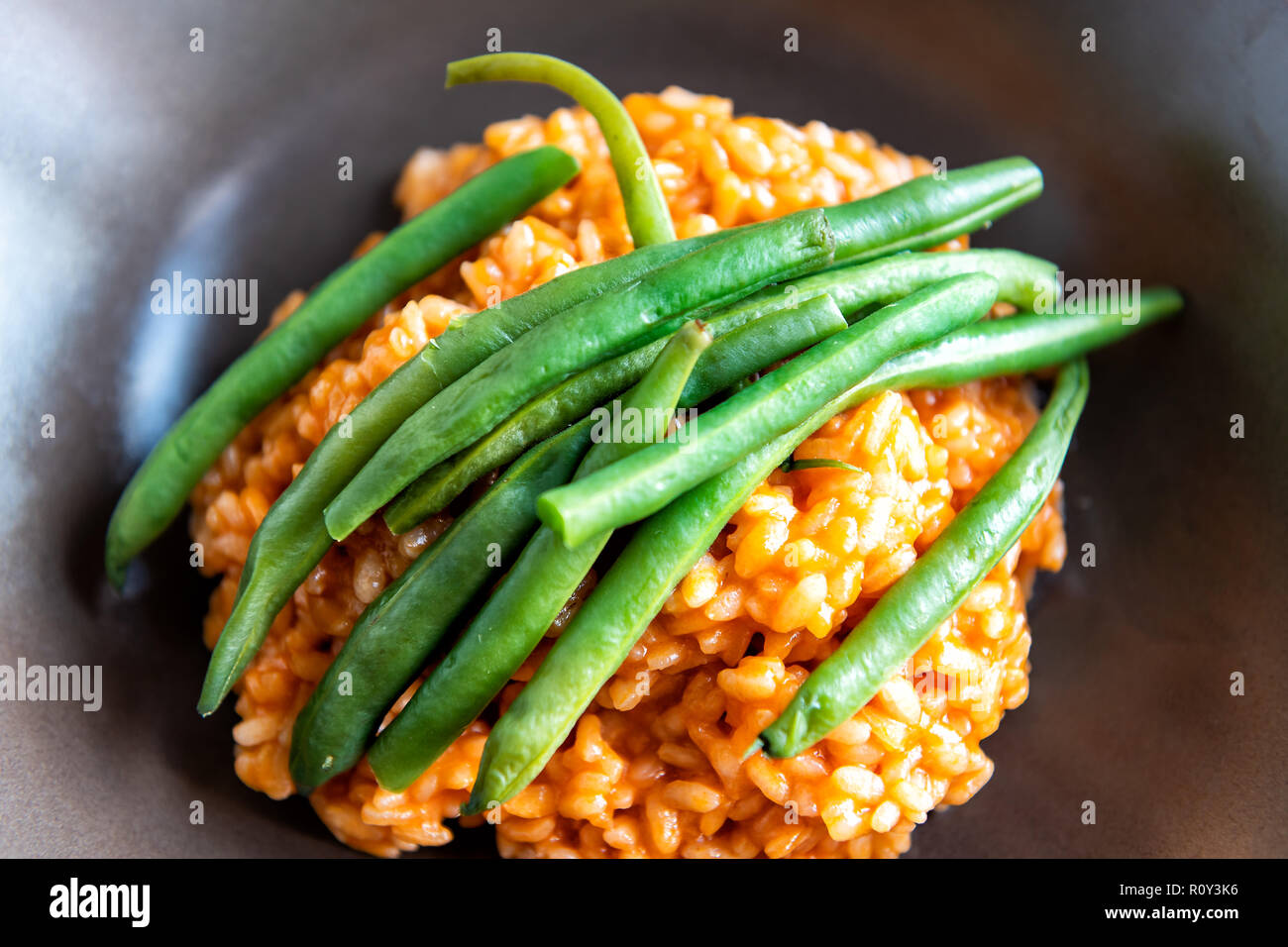Macro closeup flat top view of risotto Italian arborio cooked rice with tomato sauce, olive oil, green beans on top in brown dish, plate on table Stock Photo