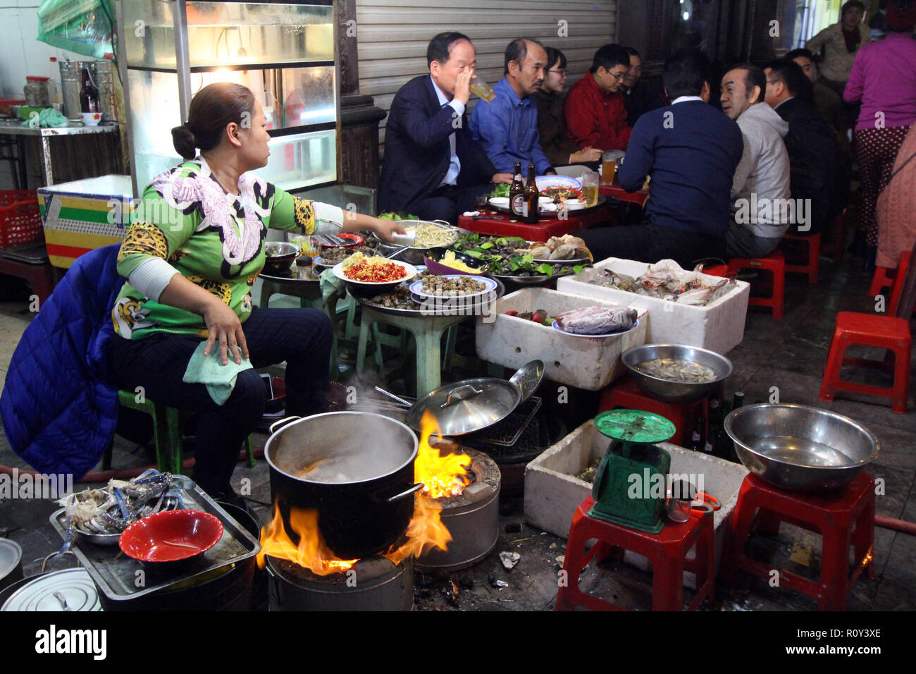 Group of men patronize a sidewalk eating establishment while the proprietor cooks over an open flame in Hanoi, Vietnam Stock Photo