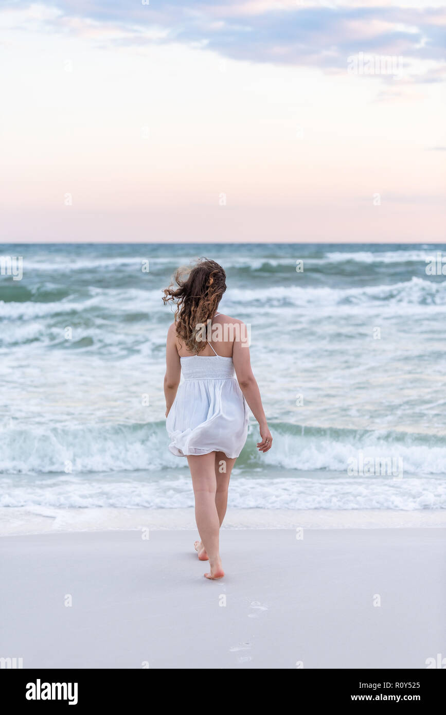 Young woman in white dress on beach pink sunset in Florida panhandle with wind, ocean waves, walking legs back to water Stock Photo