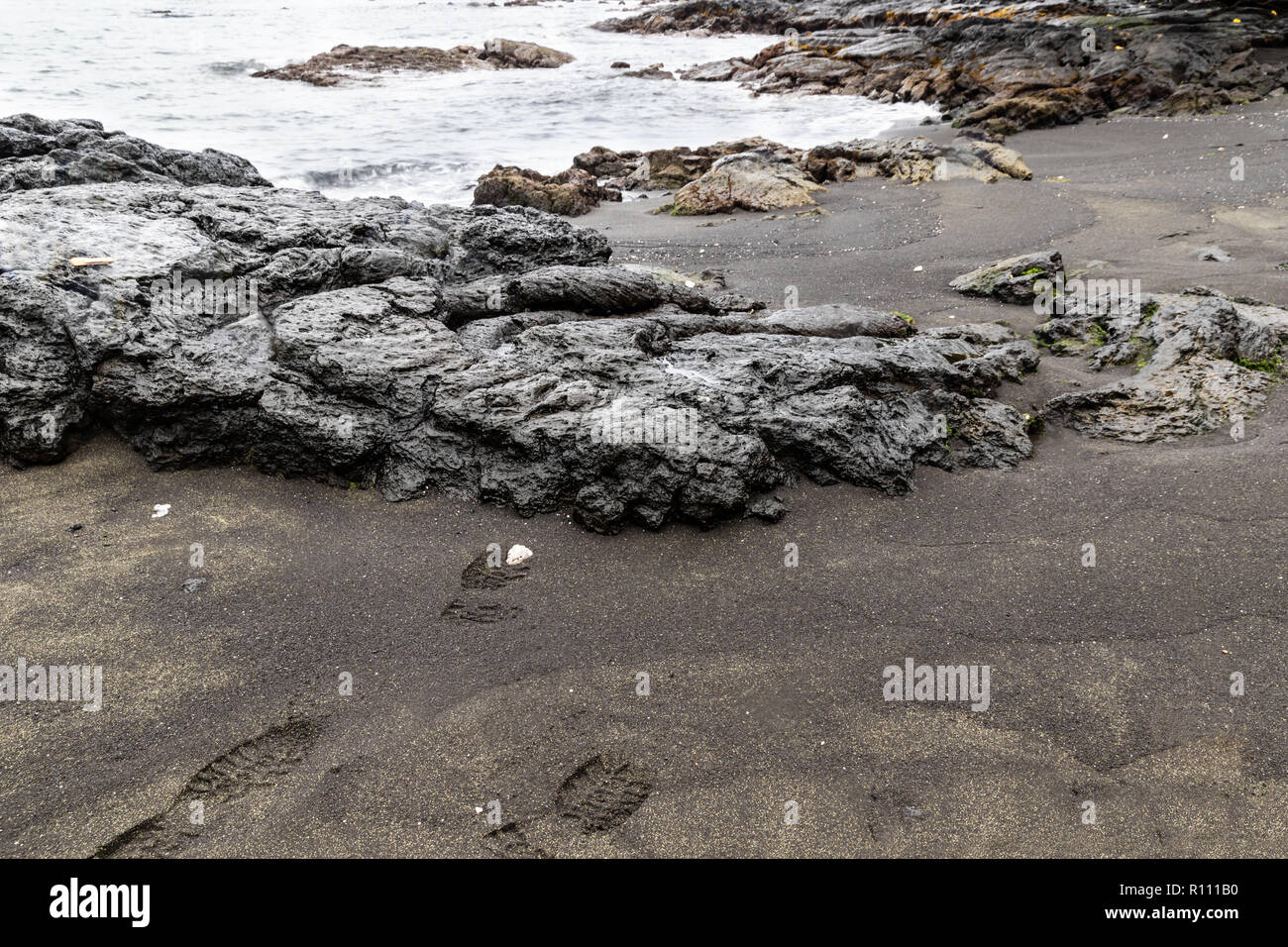 Volcanic rock on shore at green sand beach near Hilo, hawaii. Boot prints in sand, additional rocks and sea is in the background. Stock Photo