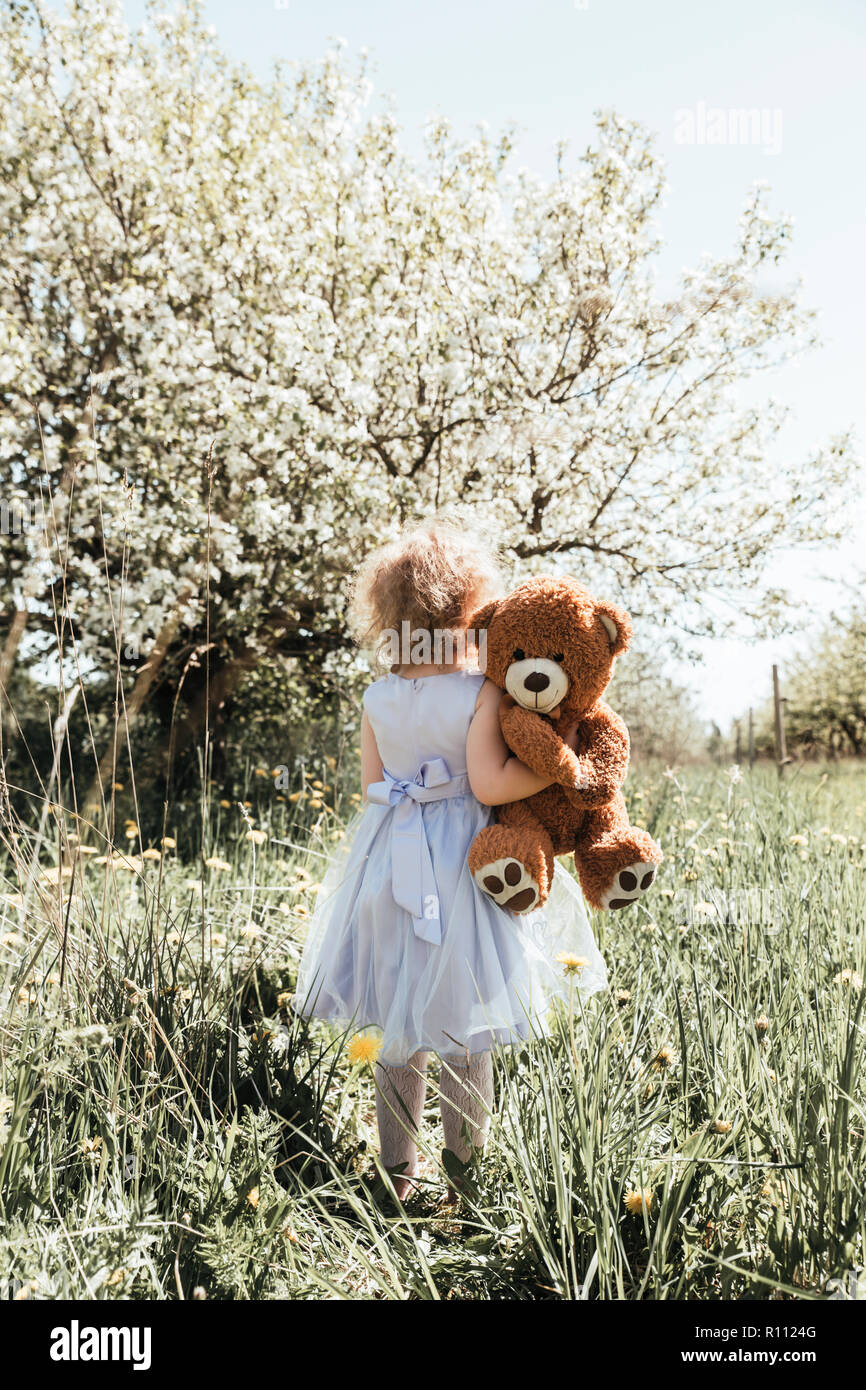 Young girl in pretty violet dress standing outdoors next to a beautiful blooming apple tree and hugging her favorite stuffed toy bear. Stock Photo