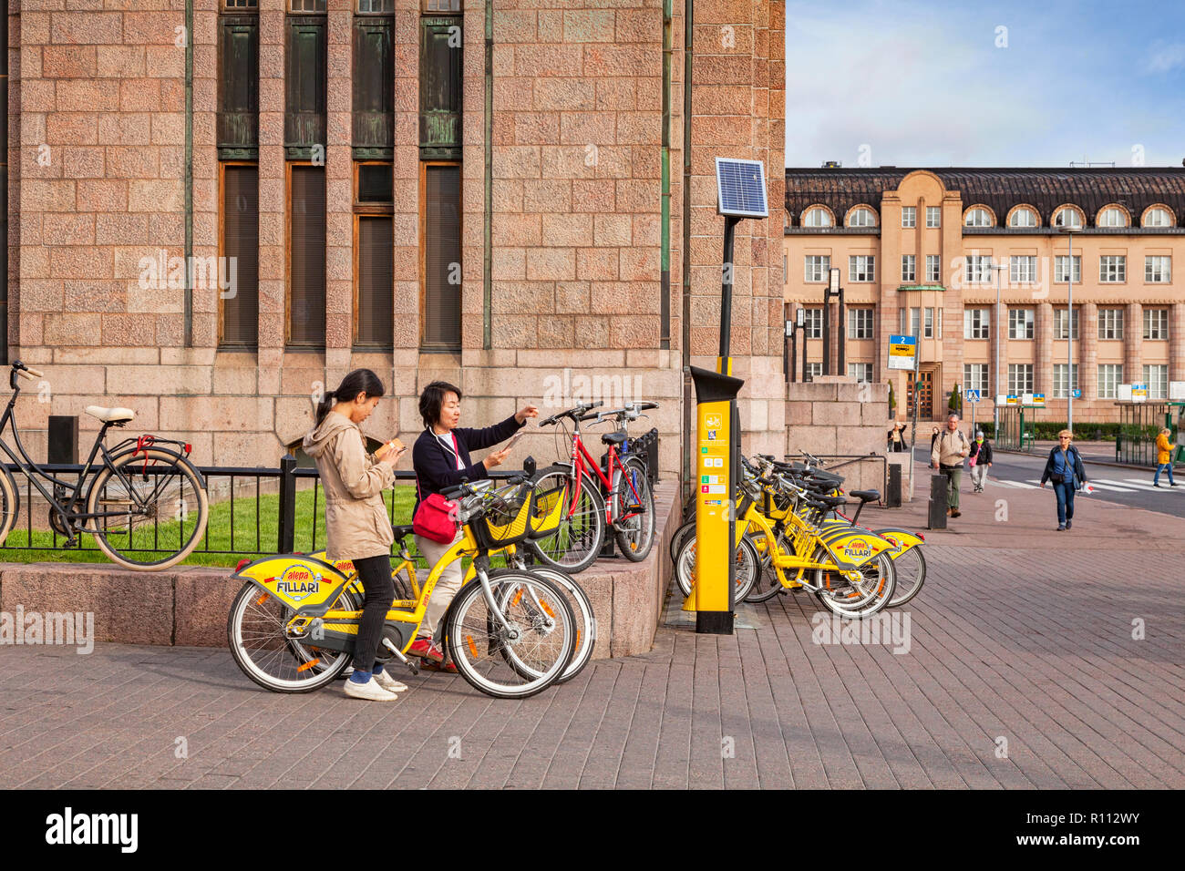 20 September 2018: Helsinki, Finland - Two female Asian tourists using yellow Alepa city bikes whilst using tablet to check map, at the central Railwa Stock Photo
