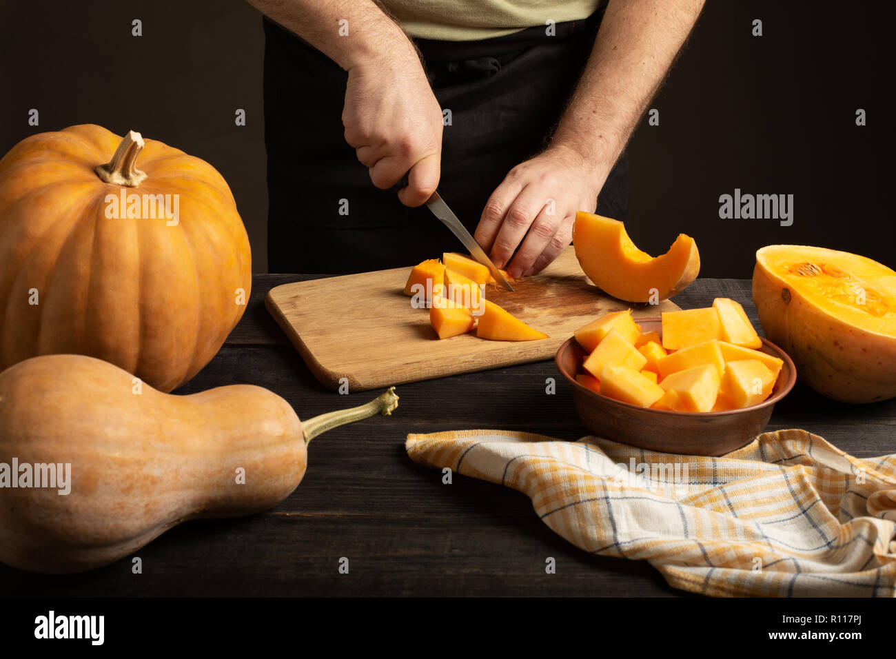 The cook slices the pumpkin for baking. On a black wooden table are whole pumpkins of different sizes. Stock Photo