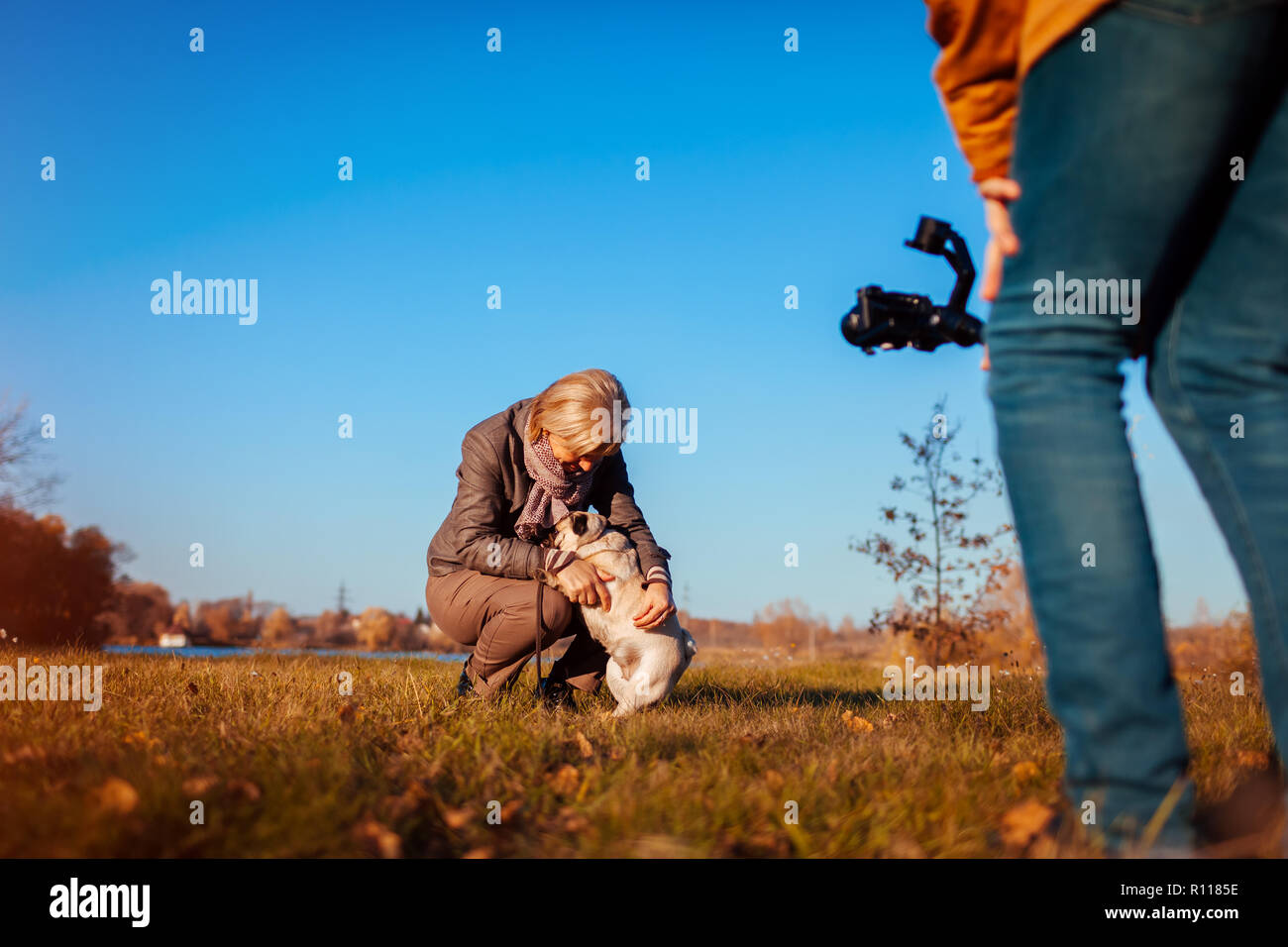 Videographer filming woman with dog in autumn park. Man using steadicam and camera to make footage. Video shoot Stock Photo