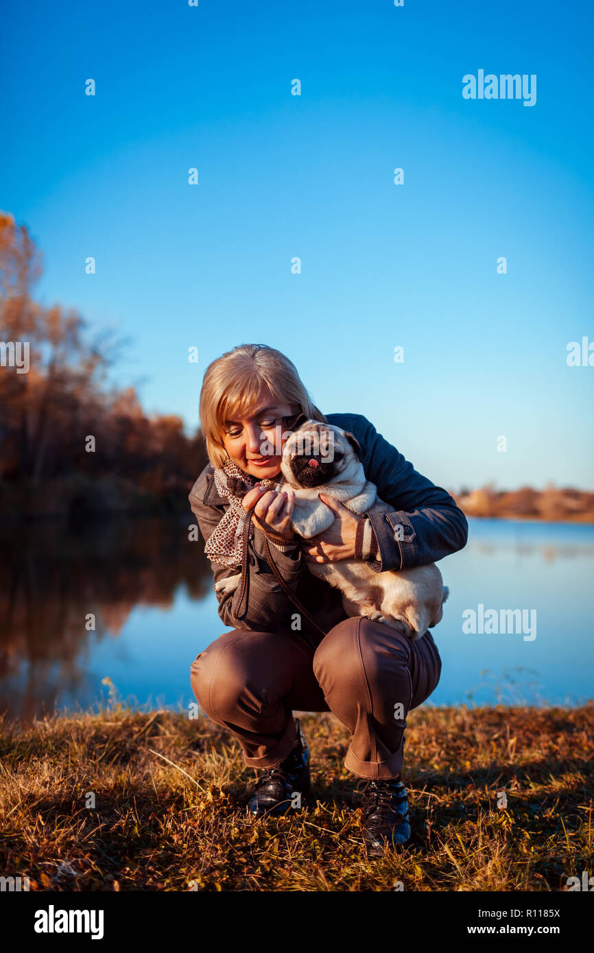 Master walking pug dog in autumn park by river. Happy woman hugging pet. Stock Photo