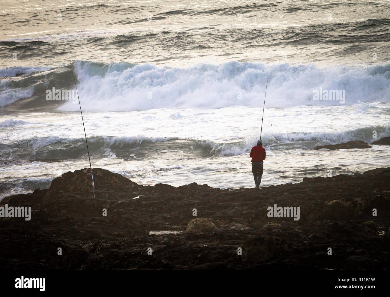 A fisherman and his fishing poles at the rocky shore, near the ocean. Waves, back view. Stock Photo