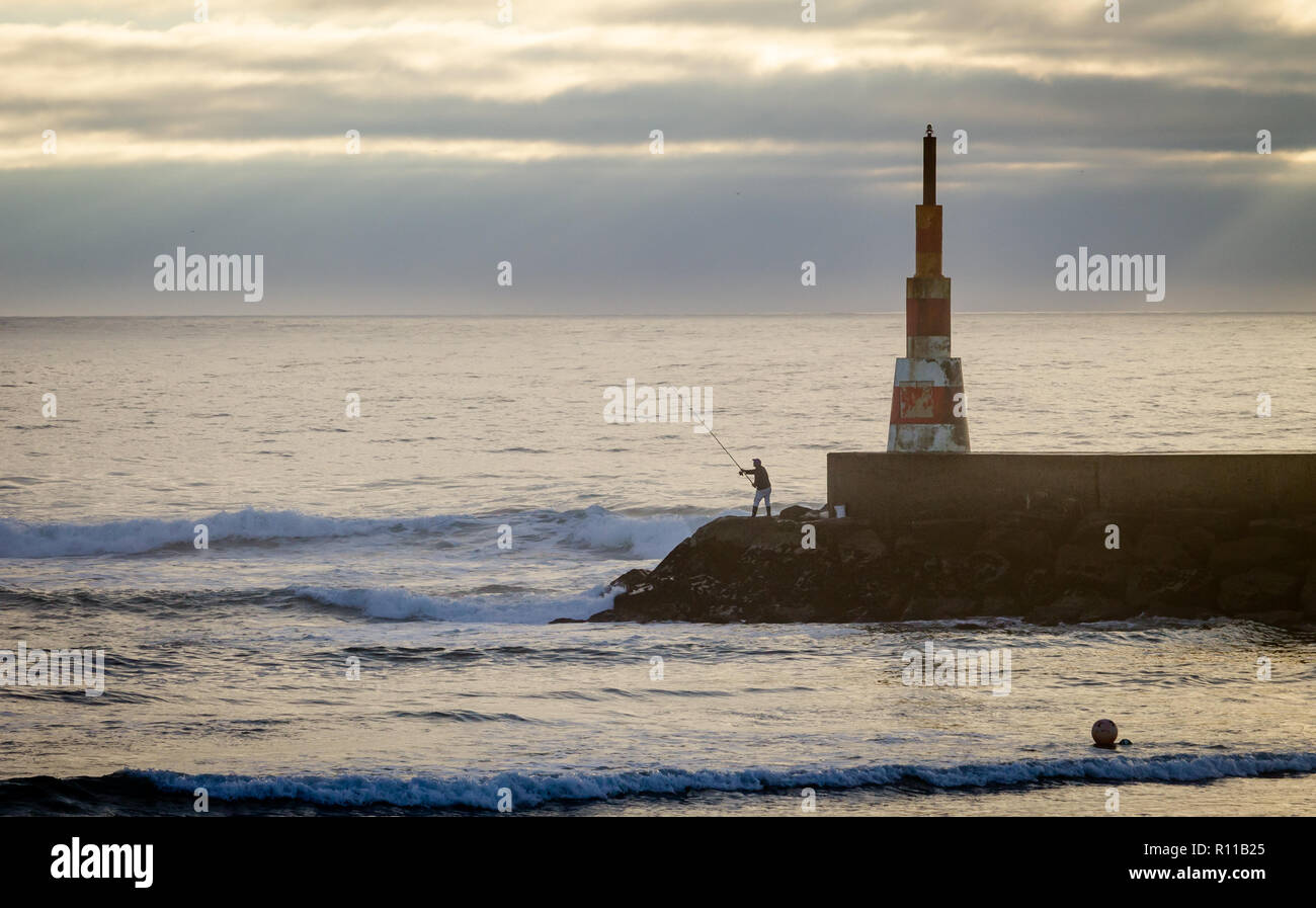 A fisherman fishes near a beacon in the ocean, cloudy day. Stock Photo