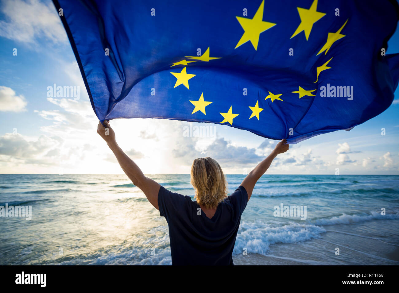 Man with blond hair standing on the shore of a Mediterranean beach holding a European Union EU flag blowing in the wind Stock Photo