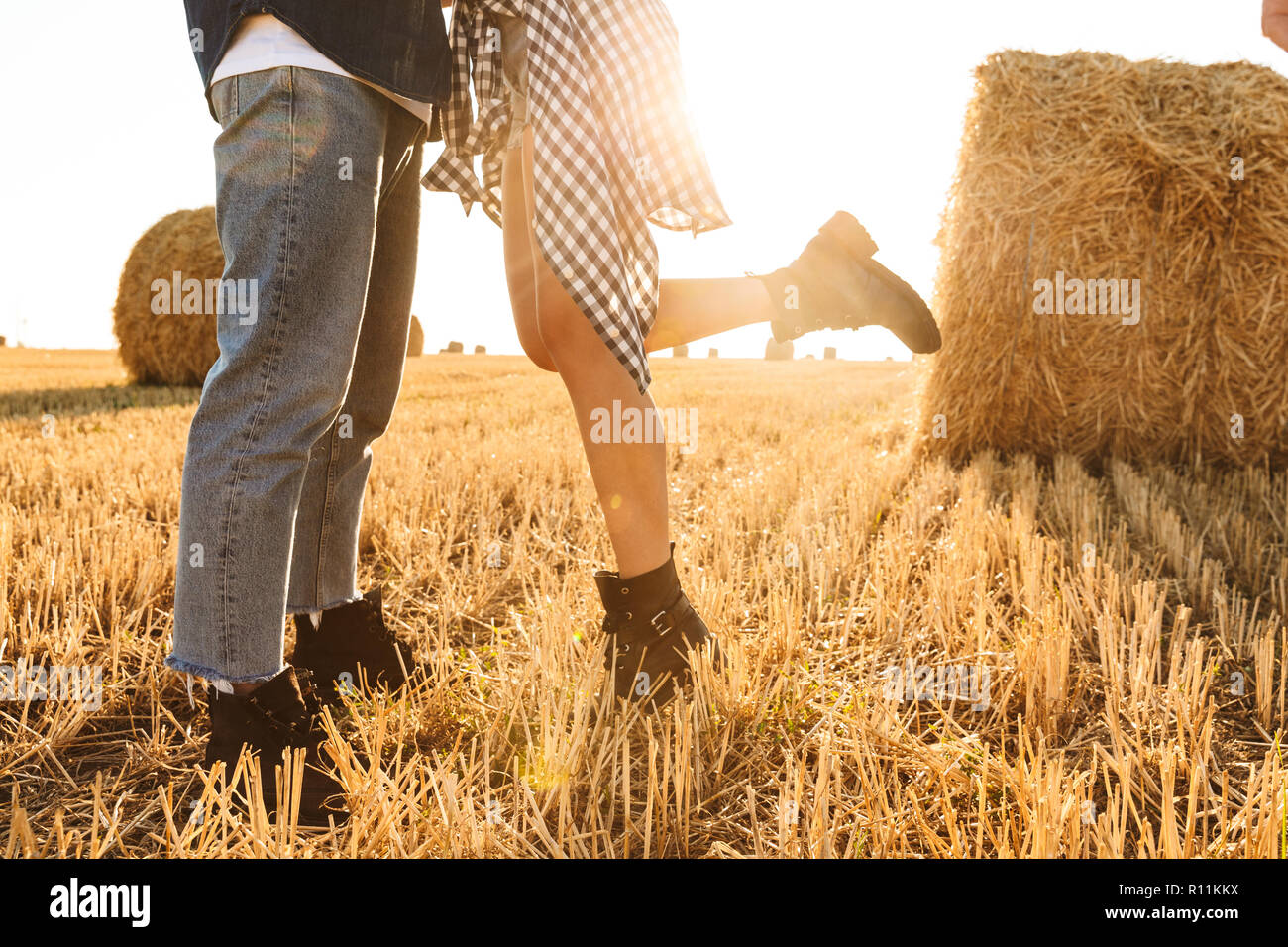 Cropped photo of guy and girl walking through golden field with bunch of haystacks and kissing during sunny day Stock Photo