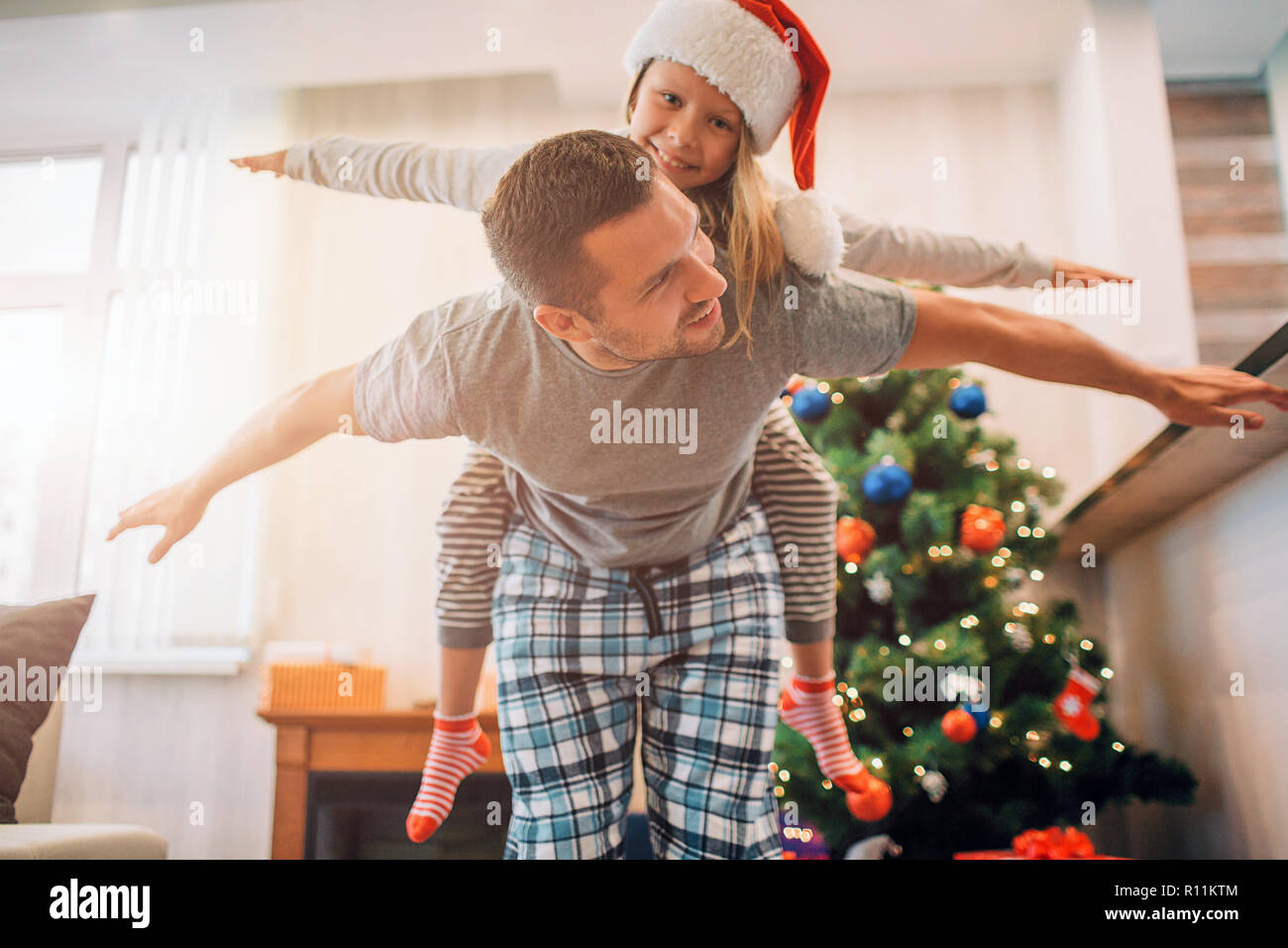 Father and daughter playing in room. He rides her on his back. They keeps hands aside of body. Young man looks at girl. They smile. People are positiv Stock Photo