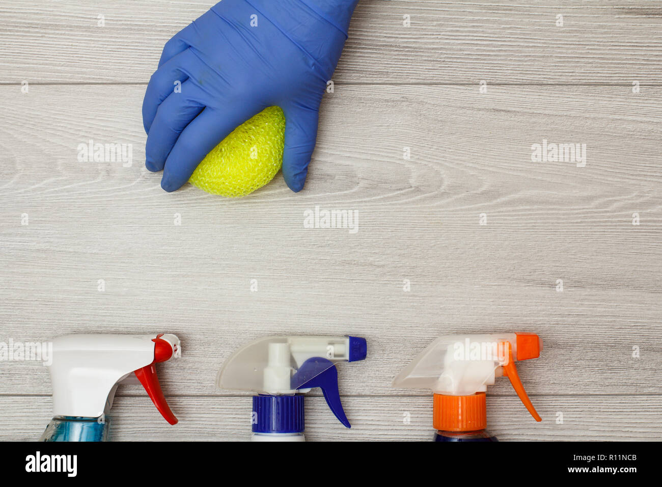 Hand in rubber glove holding synthetic sponge for cleaning with bottles of detergent on wooden boards. Top view Stock Photo