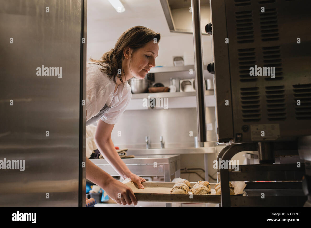 Mid adult female baker placing a tray of uncooked scones into the oven. Stock Photo