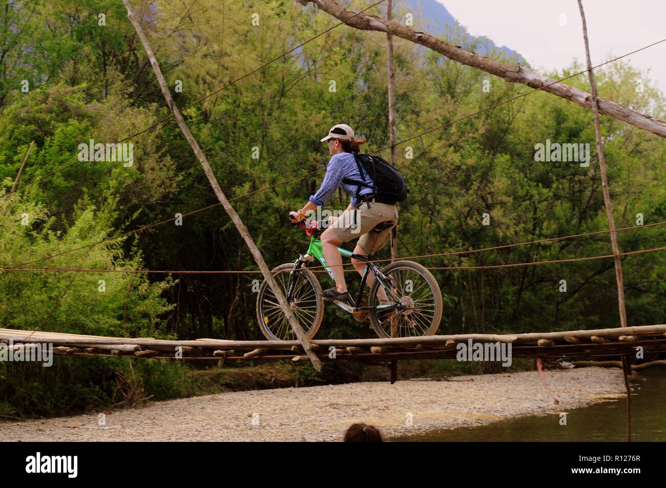Sporty woman rides a bicycle on a wooden bridge across the river Nom Song, Vang Vieng, Laos. Stock Photo