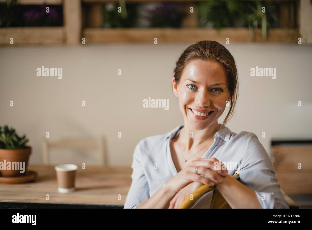 Headshot of a mid adult woman sitting indoors in a small coffee shop. She is looking at the camera and smiling. Stock Photo