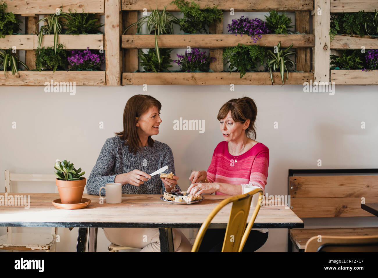 Two Female friends sitting inside a little cafe relaxing. They are buttering a scone and enjoying a coffee. Stock Photo