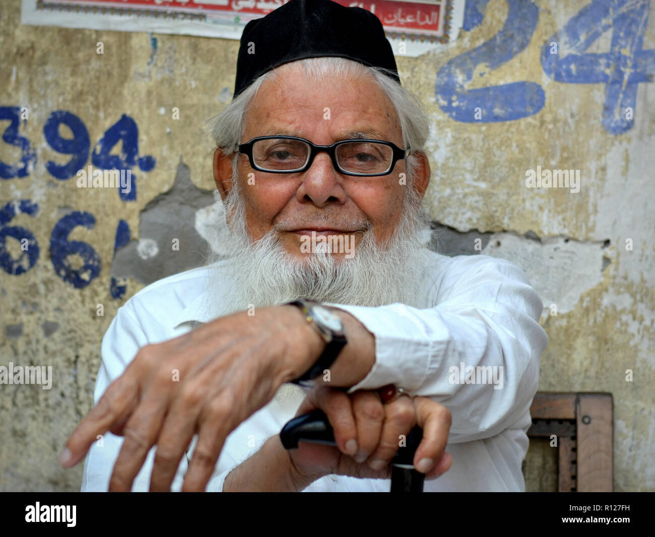 Sitting elderly Indian Muslim man with a white Islamic beard wears his traditional white Islamic clothing and a black skull cap (taqiyah). Stock Photo