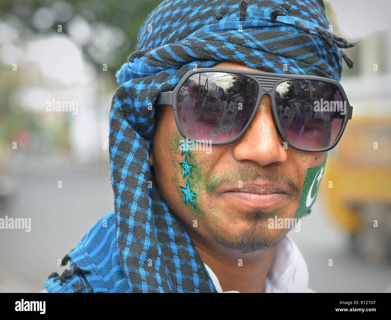 Young Indian Muslim man with green glitter and an Islamic flag sticker on his cheeks wears fancy aviator-style sunglasses and a checkered headscarf. Stock Photo