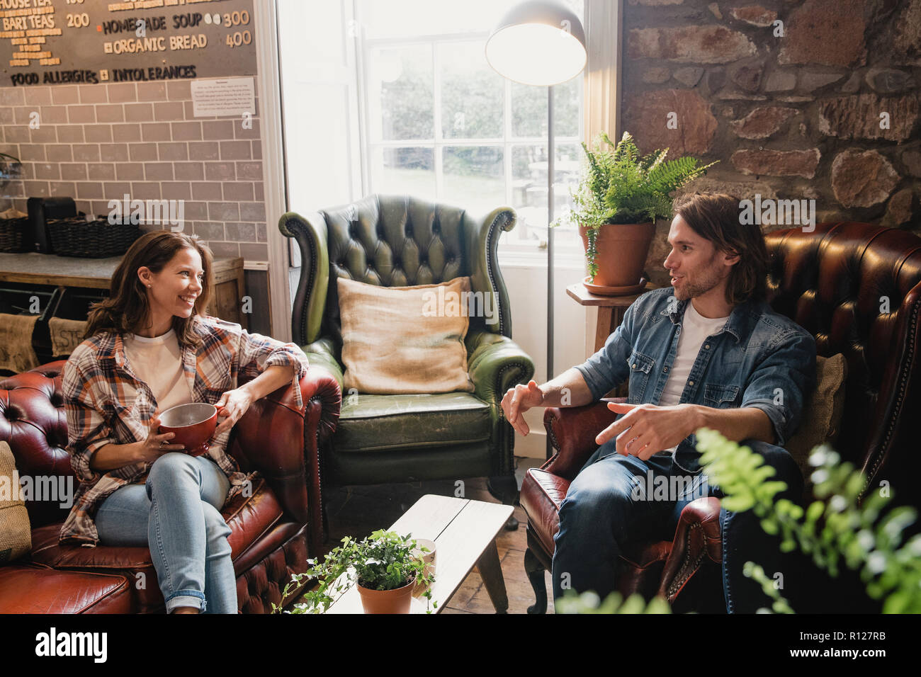 Mid-adult couple sitting in a small coffee shop relaxing. They are sitting on leather sofas and enjoying a coffee while talking. Stock Photo
