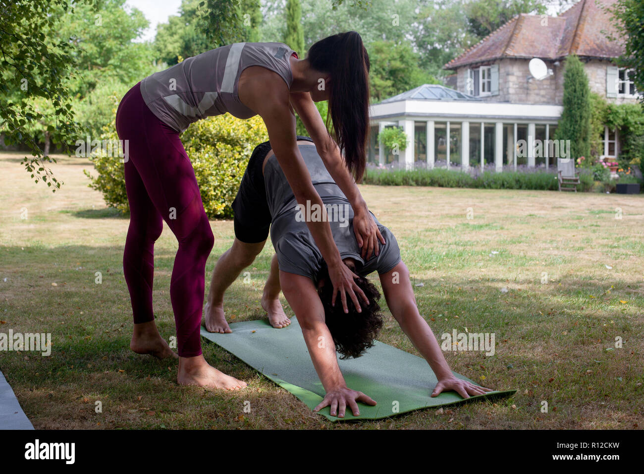 Female yoga teacher teaching young man yoga in garden, downward facing dog pose Stock Photo