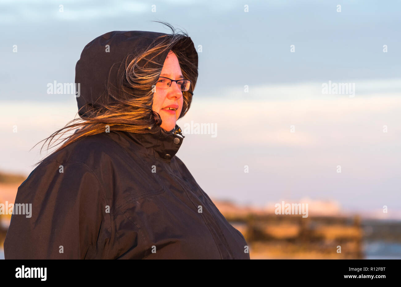 Woman looking to be in her 30's standing outside in a coat with hood up watching the sun go down with sunlight shining on her face, in the UK. Stock Photo