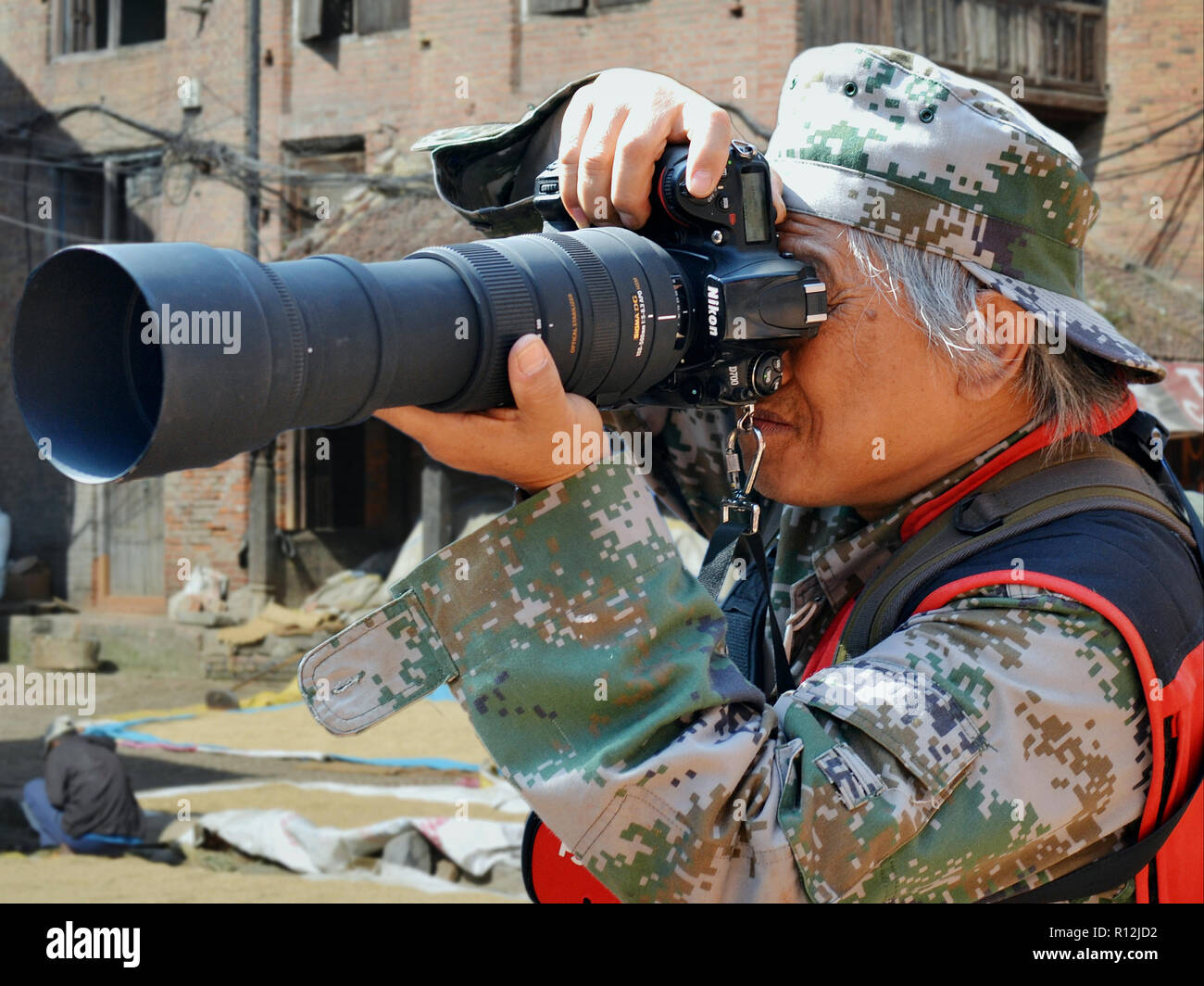 Elderly, camouflaged Chinese street photographer takes a photo with a Nikon DSLR camera with high-power telephoto lens. Stock Photo
