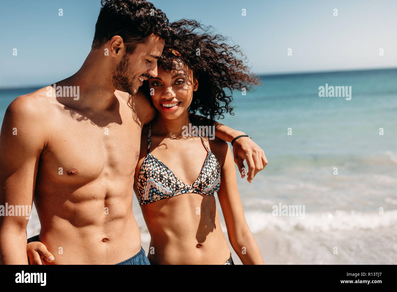 Romantic couple walking on beach together on a sunny day. Smiling man walking on beach with girlfriend putting his arm around her shoulder. Stock Photo