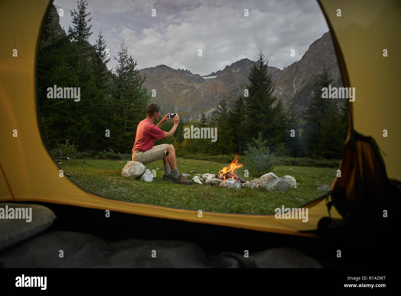View from inside tourist tent. Man hiker sitting at bonfire, making selfie photo by mobile phone Stock Photo