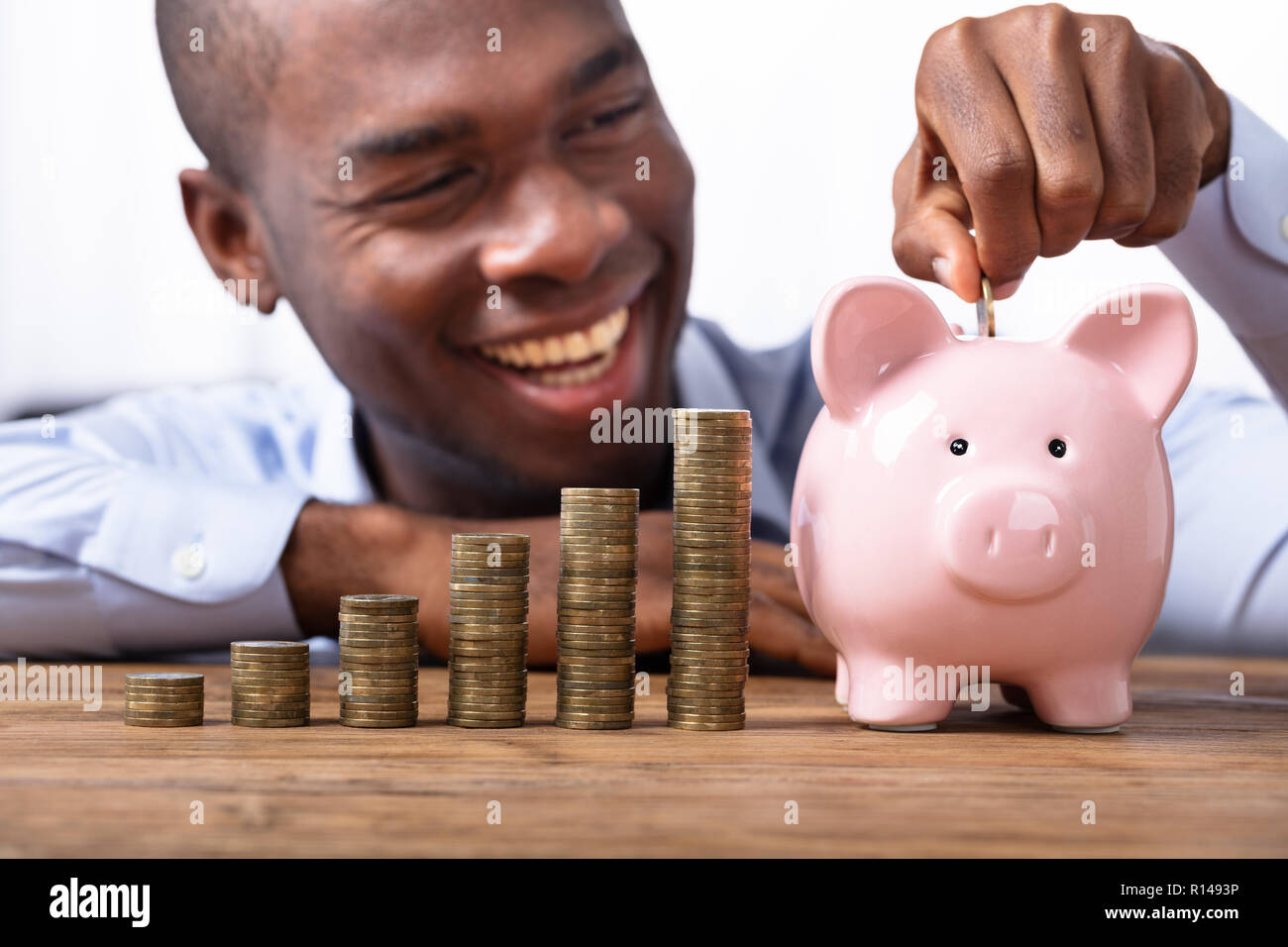 Happy African Man Inserting Coin In Piggybank Near Stack Of Coins Stock ...