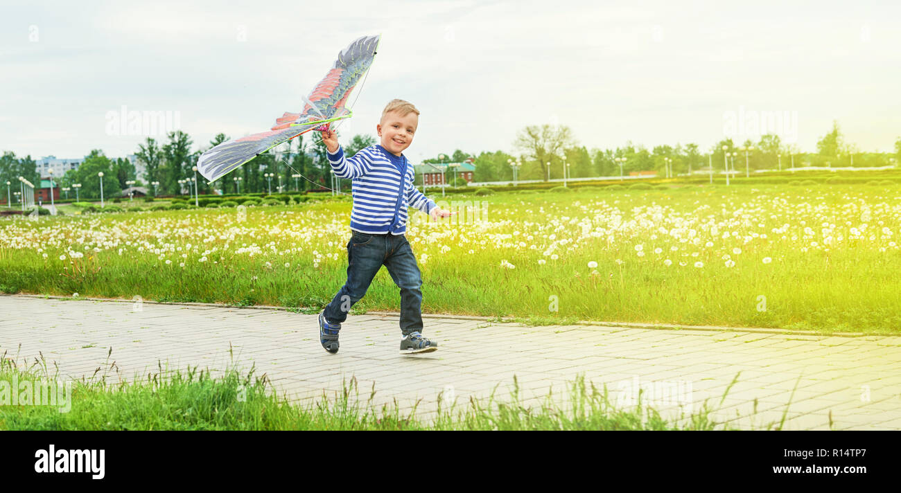 Little boy running across park with toy kite flying. Caucasian child playing on spring day. Lifestyle kid actively recreation .Copy space for text,blank background. Summer,outdoors, green grass. Stock Photo