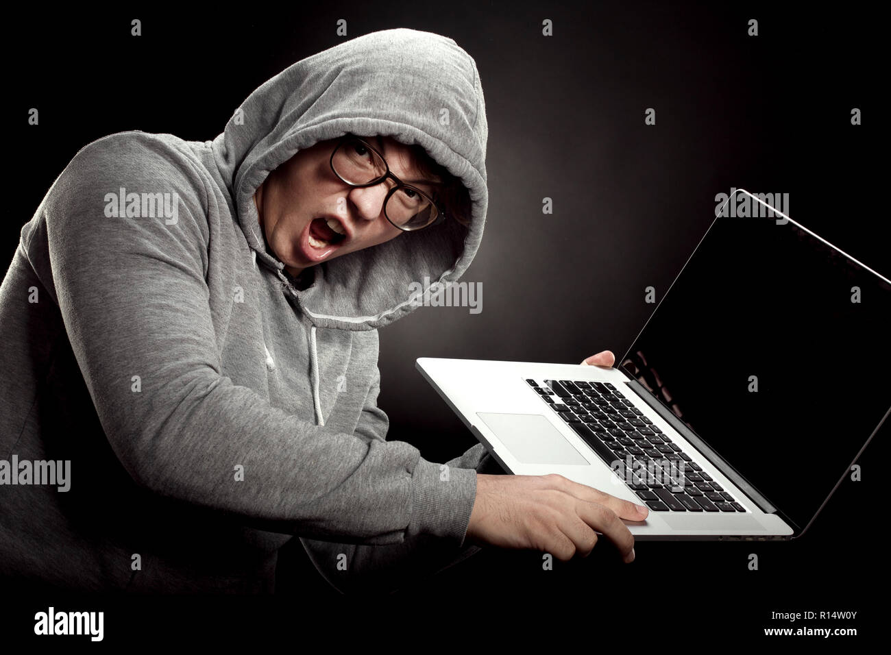 a programmer in the hood and glasses screaming furiously and is about to throw a laptop at the wall Stock Photo