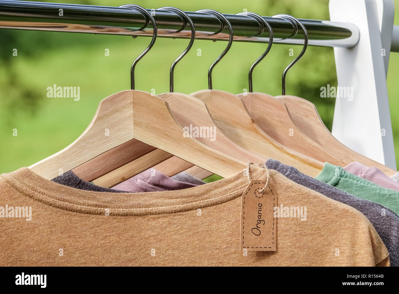 Organic clothes. Natural colored t-shirts hanging on wooden hangers in a row. Eco textile tag. Green forest, nature in background. Stock Photo