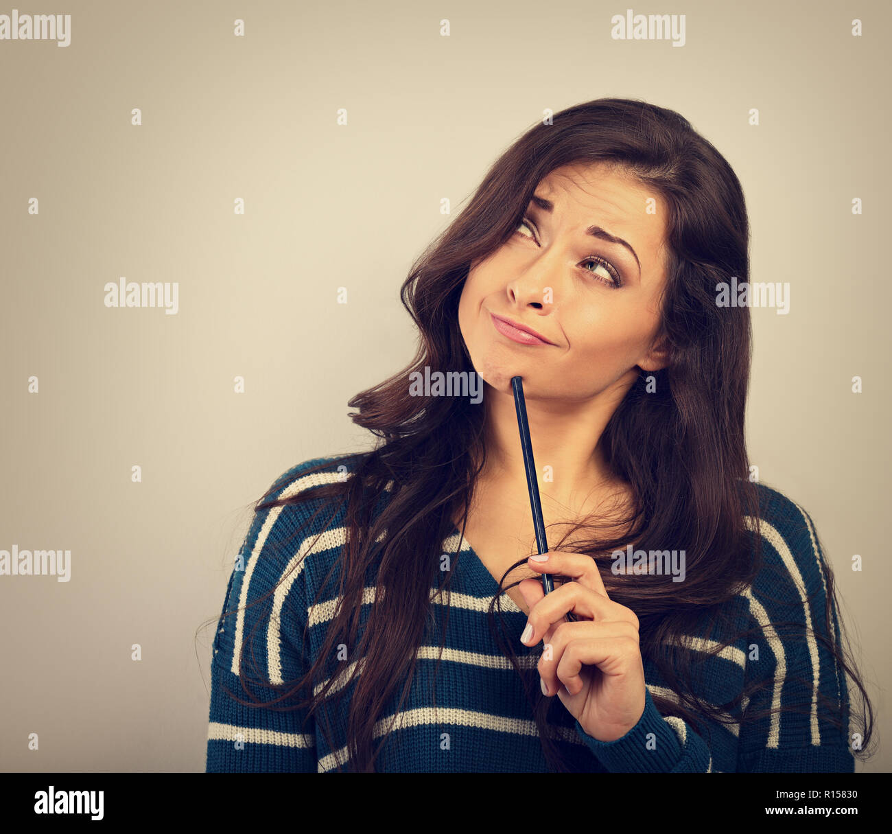 Confusion grimacing brunette woman thinking and looking up with question in eyes and pencil under the face in warm sweater. Toned closup portrait Stock Photo