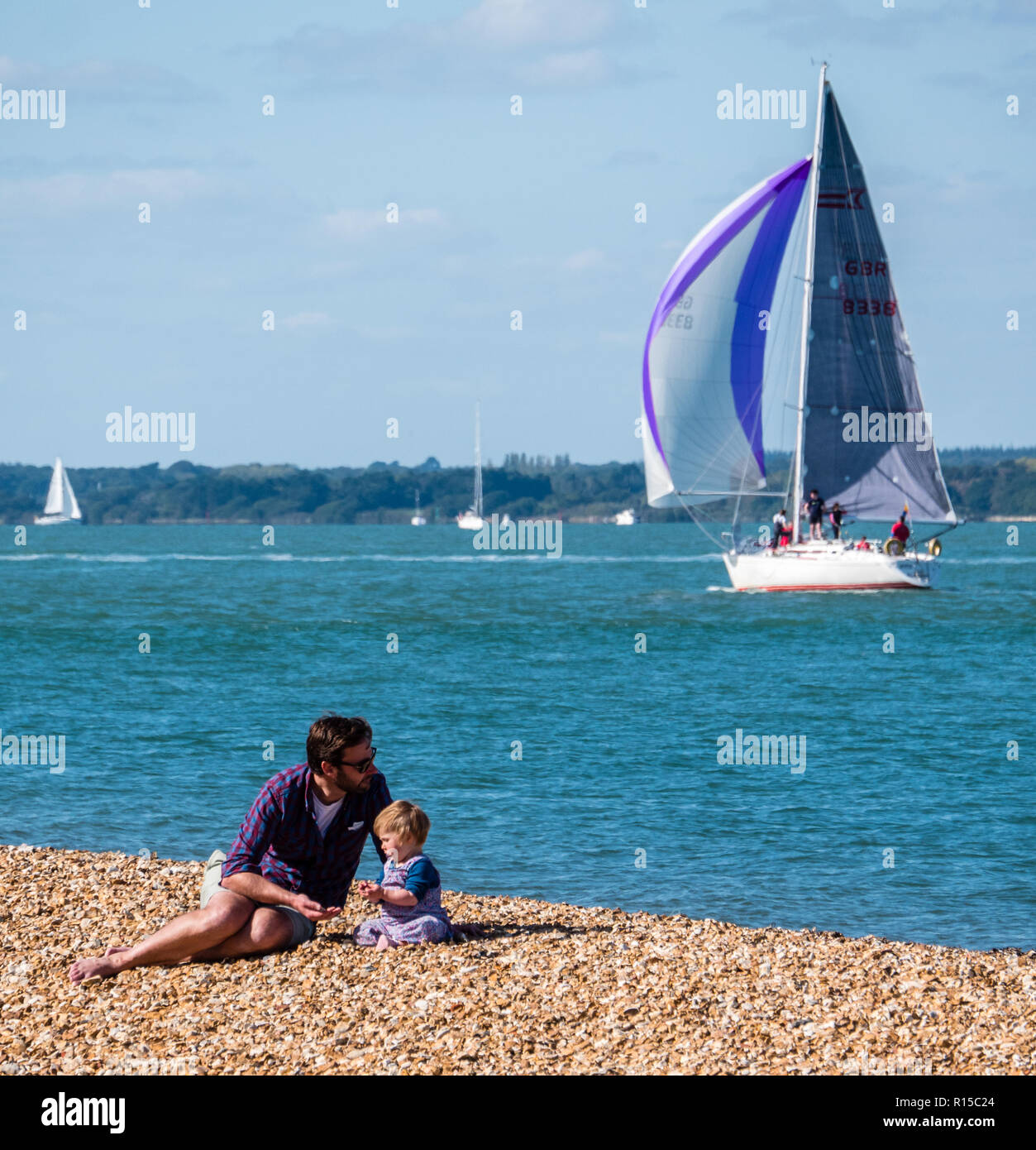 Father and Son Relaxing on Cowes, Pebble Beach, with Sailing Yacht on
