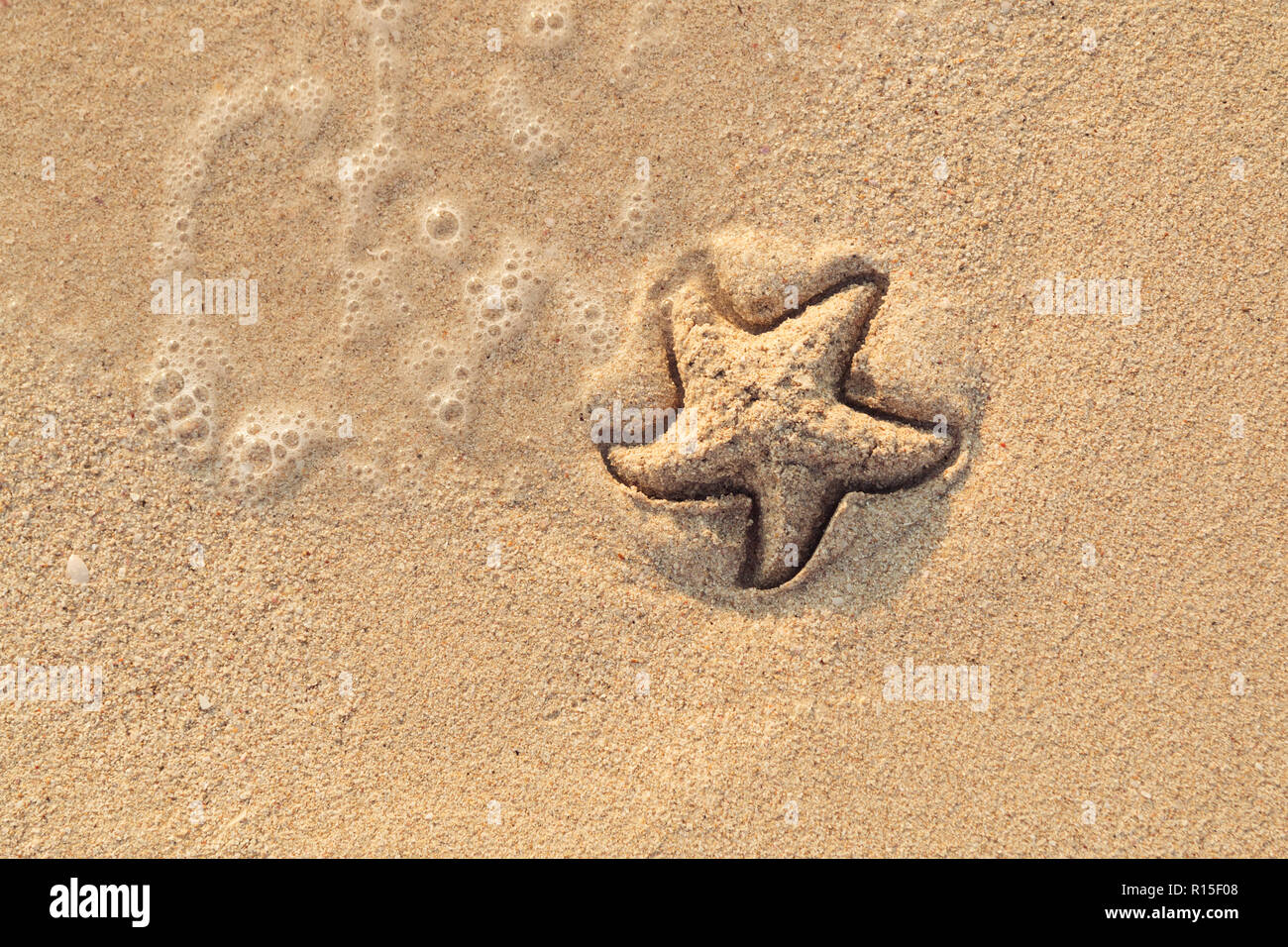 Starfish drawn on the beach sand being washed away by a wave. Foaming sea wave coming to wash a picture on wet yellow beach sand. Holiday and vocation Stock Photo