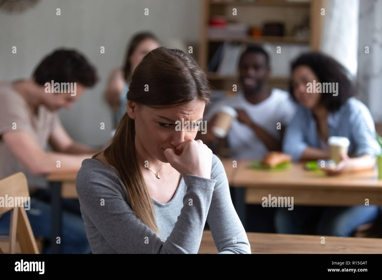 Diverse multi-ethnic friends sitting together in cafe talking having fun, focus on frustrated shy girl sitting separately by others teenagers feels un Stock Photo